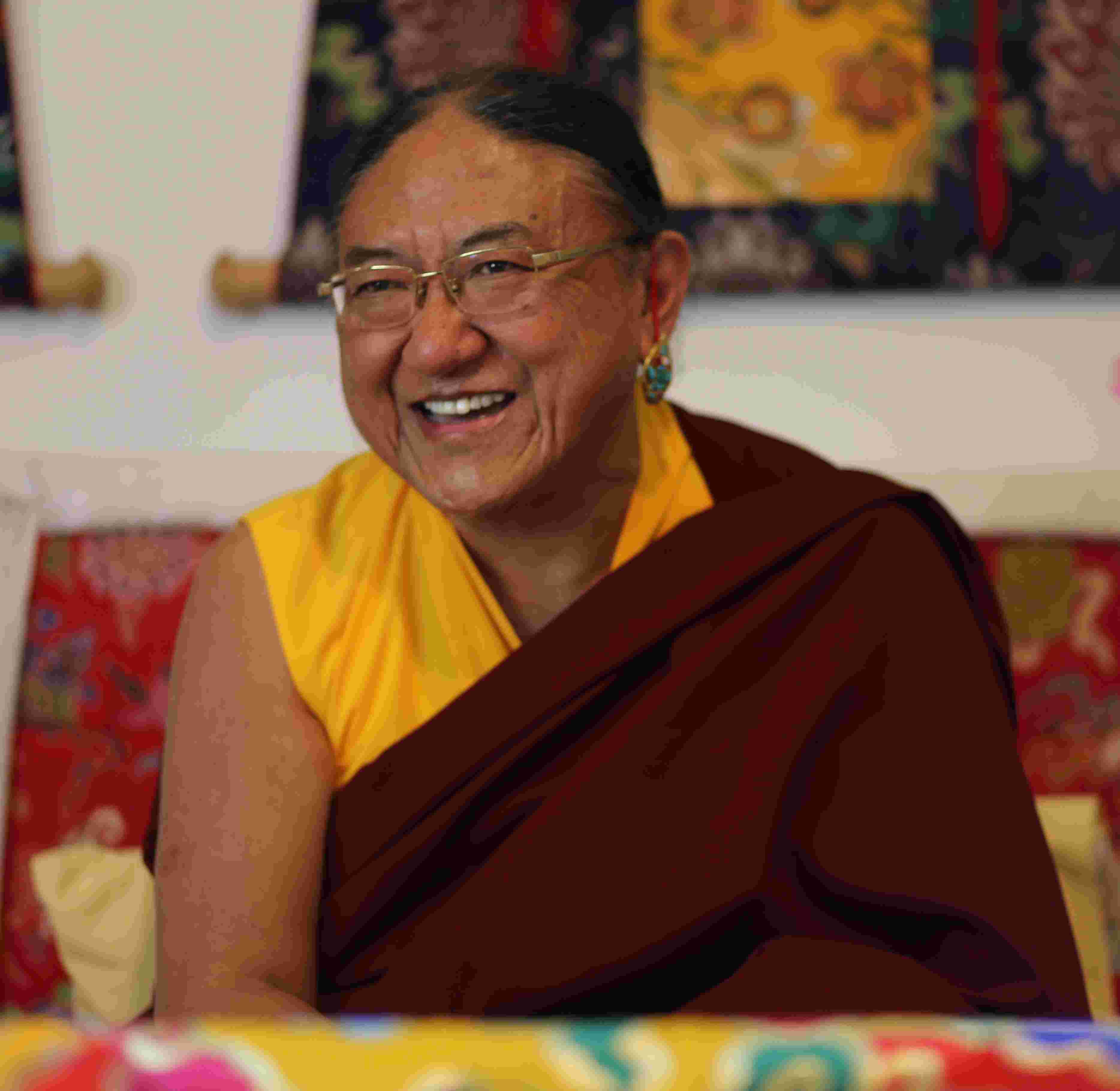His Holiness Sakya Trizin Ngawang Kunga smiling warmly in traditional maroon and saffron monastic robes, with ornate thangka paintings visible in the background